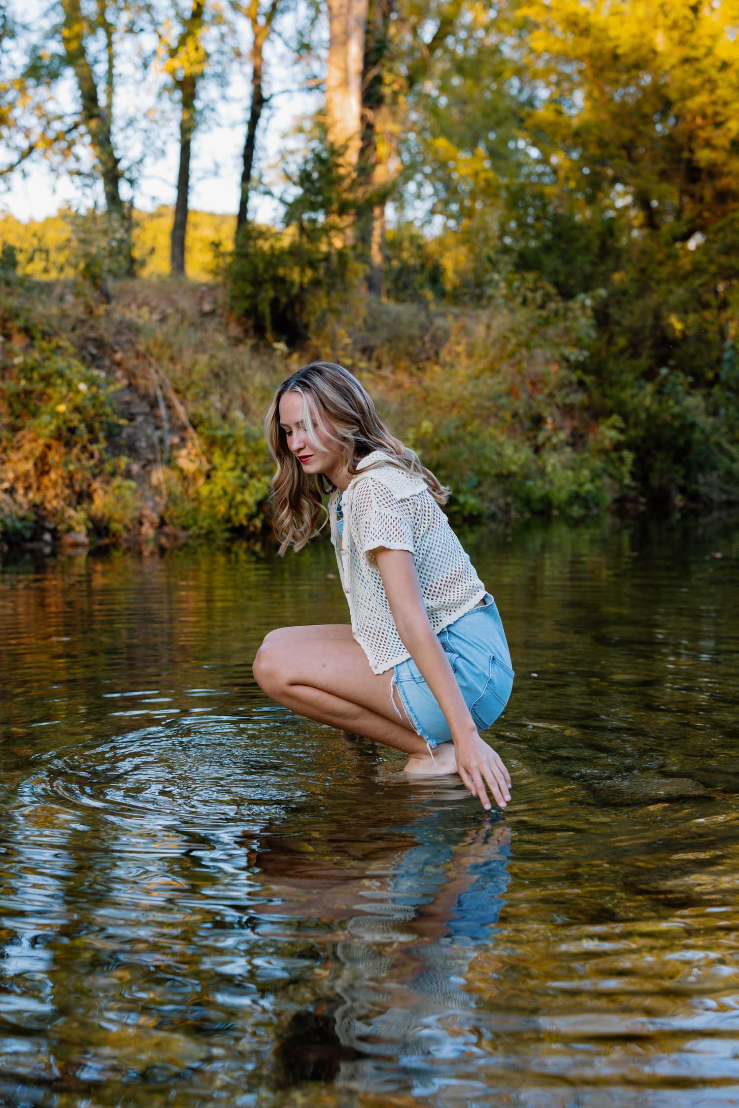 Woman in casual attire splashing in a lake during golden hour with autumn foliage in background.