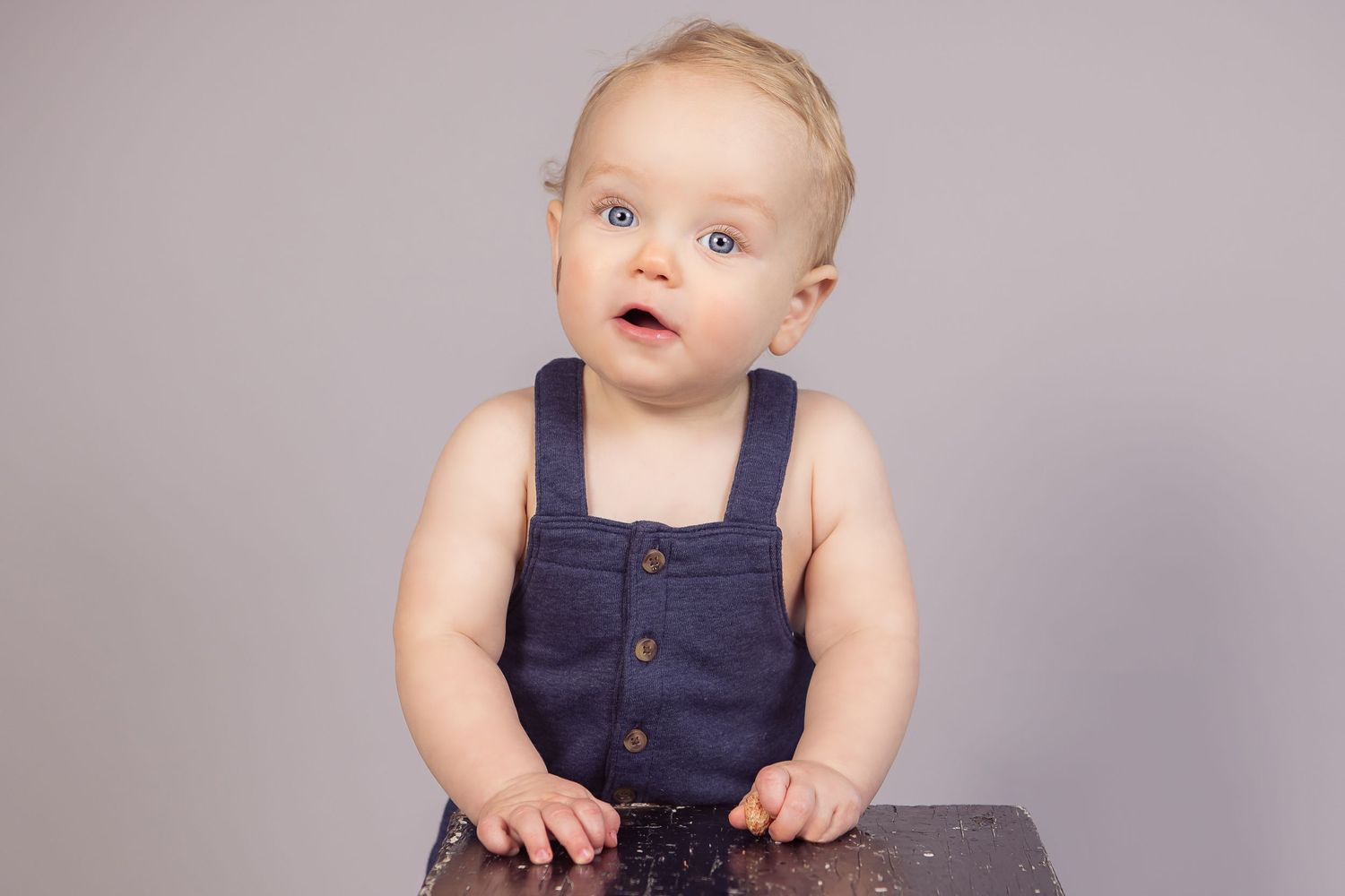 Baby in navy blue overalls sits on wooden surface against gray backdrop.