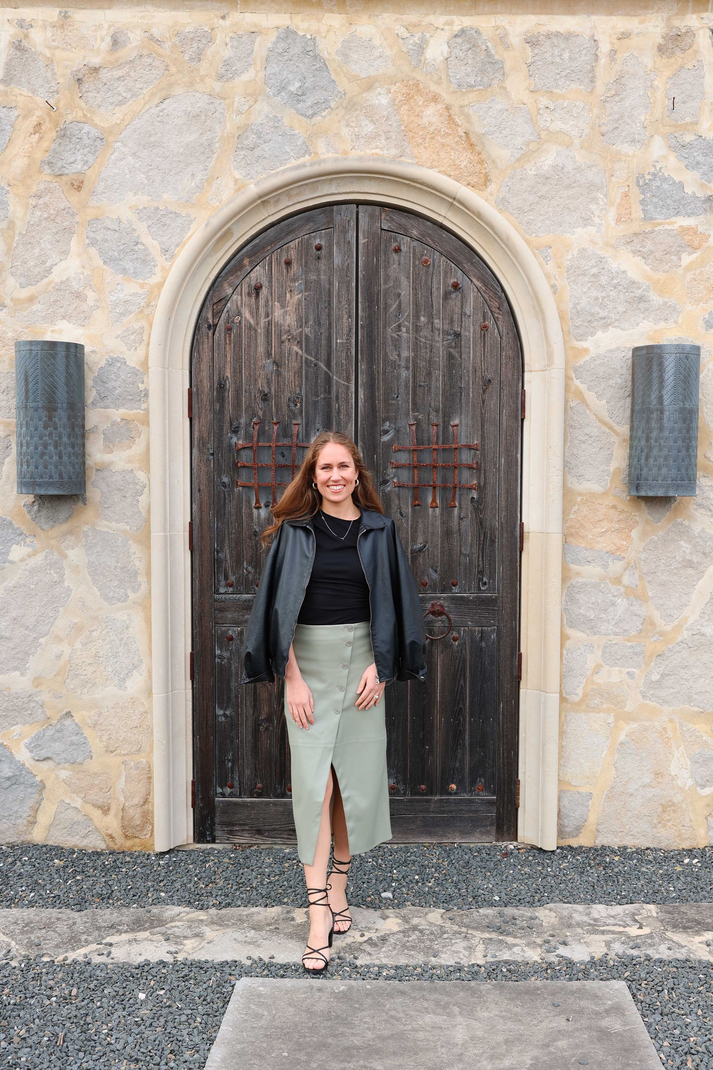 Person standing in front of arched wooden door wearing leather jacket and midi skirt.
