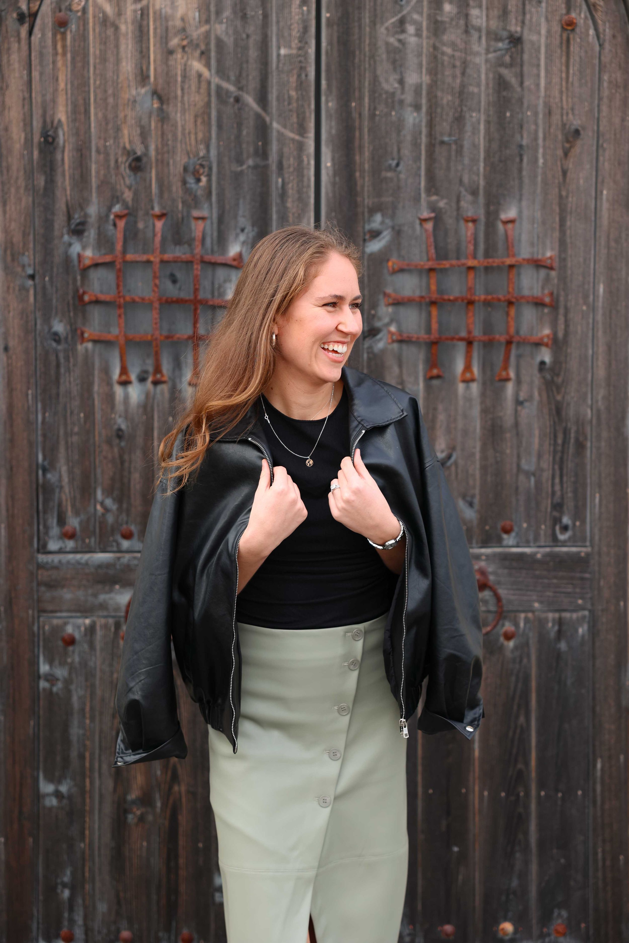 Person in black jacket and sage skirt smiling against weathered wooden door background.