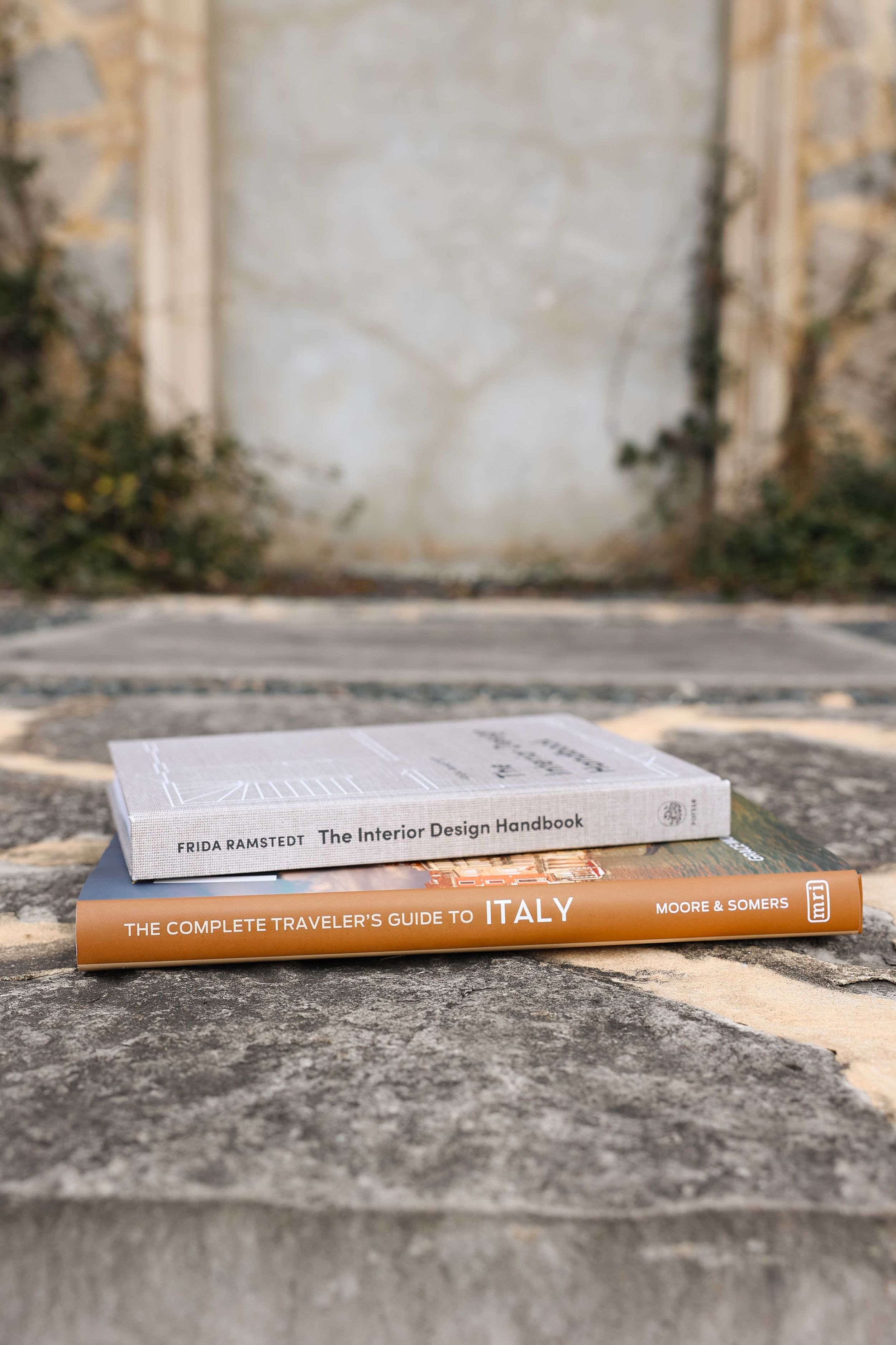 Two books about interior design and Italy lying on stone surface with blurred background.