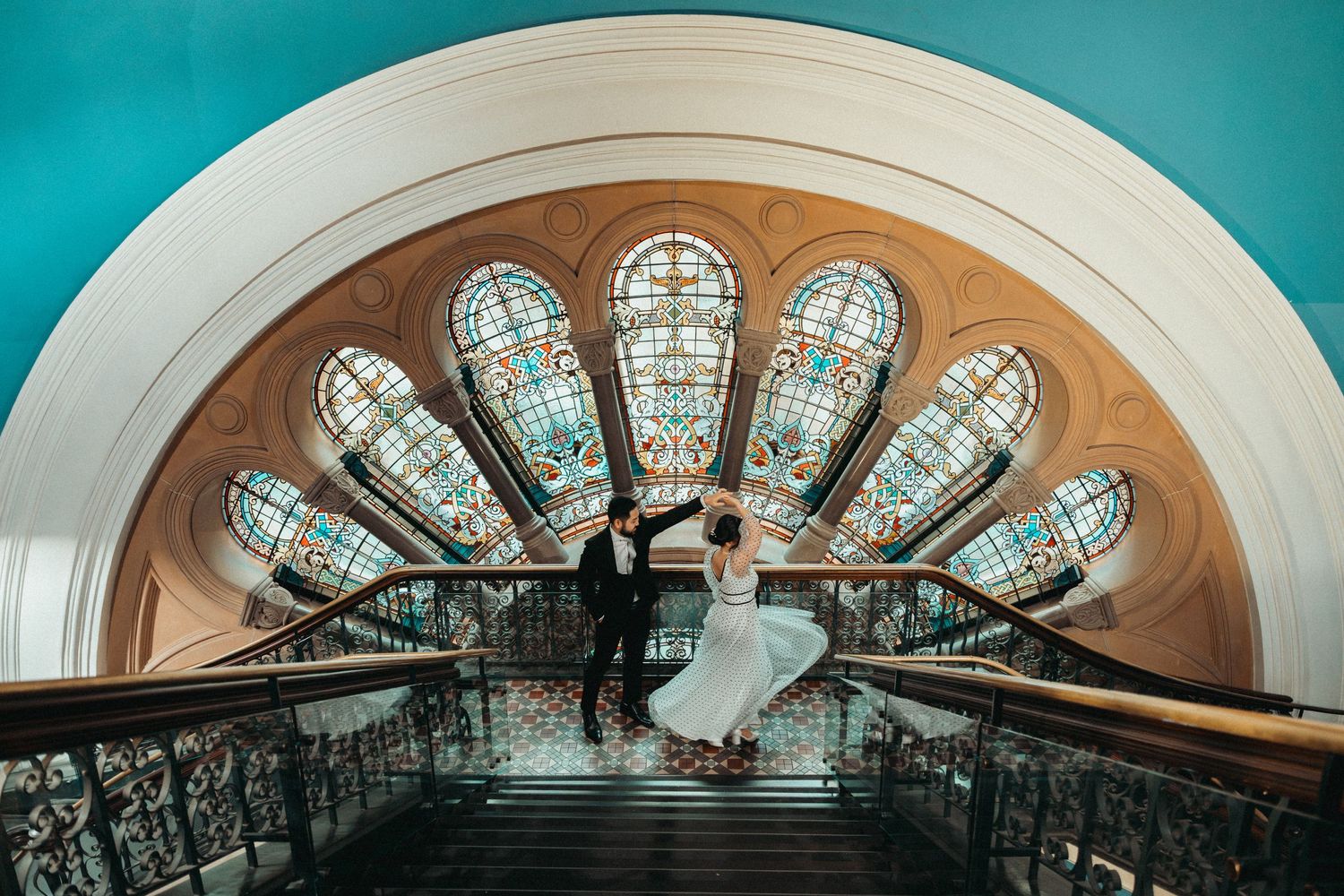Silhouetted figures stand beneath a stunning stained glass fan window in a dramatic architectural setting.