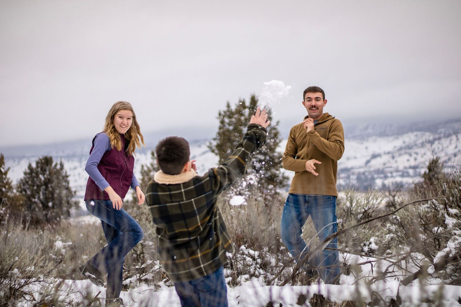 a kid throws a snowball at his brother and sister who laugh and look on. they are surrounded by snowy brush and hills
