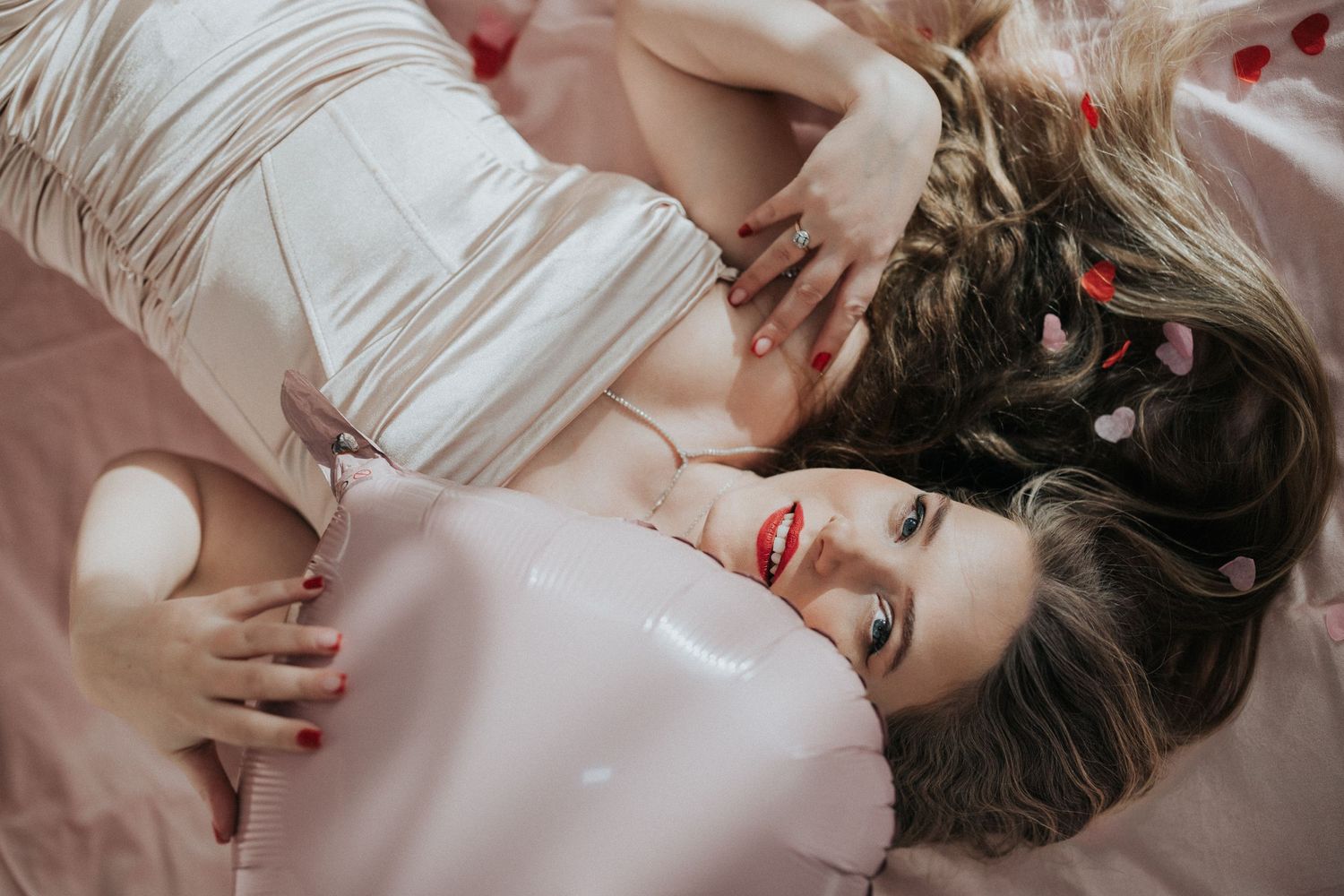 Woman with red lips lying on pink bedding surrounded by heart decorations in Valentine's shoot.