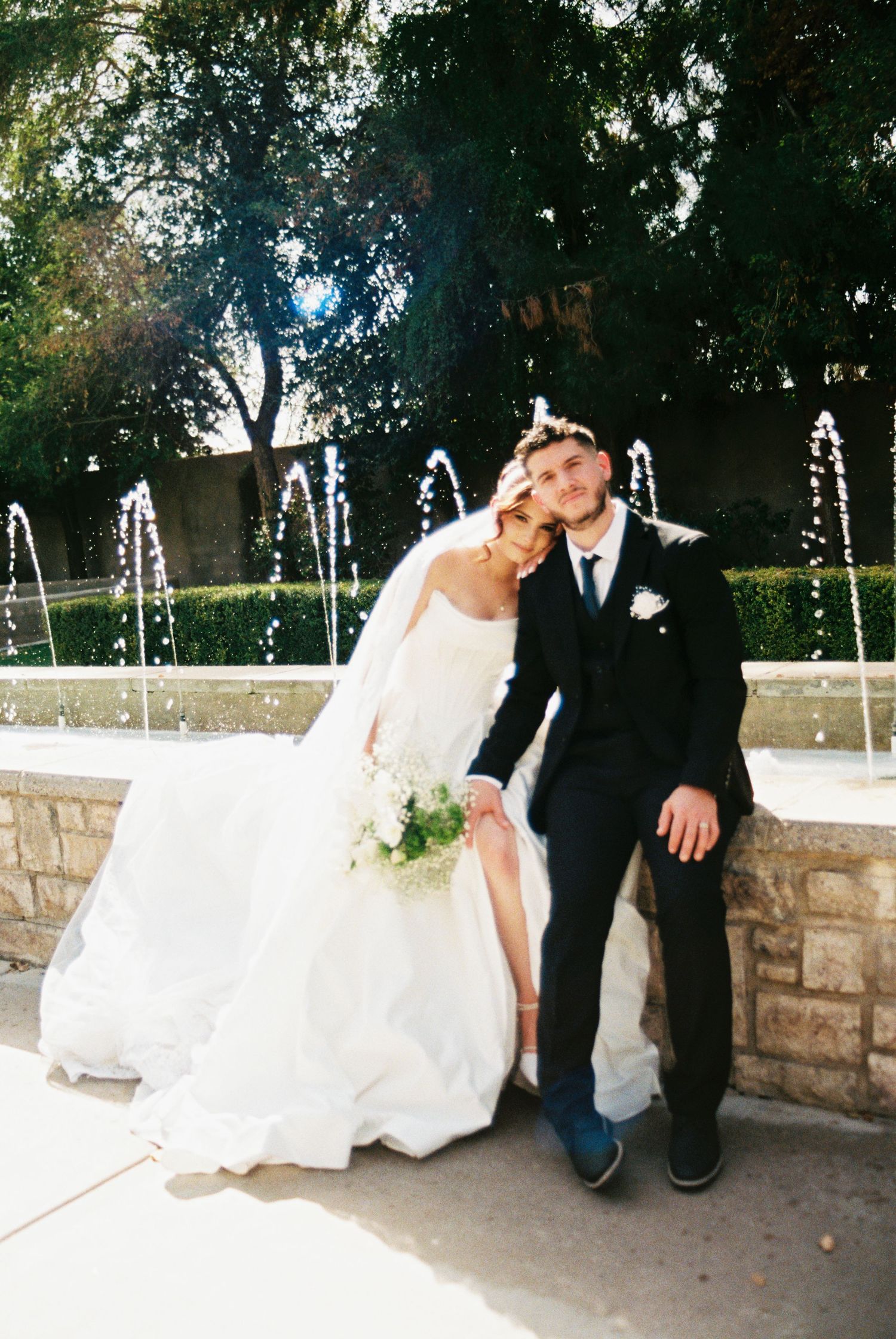Newlywed couple sits together on stone wall with decorative water fountains creating romantic ambiance in garden setting.