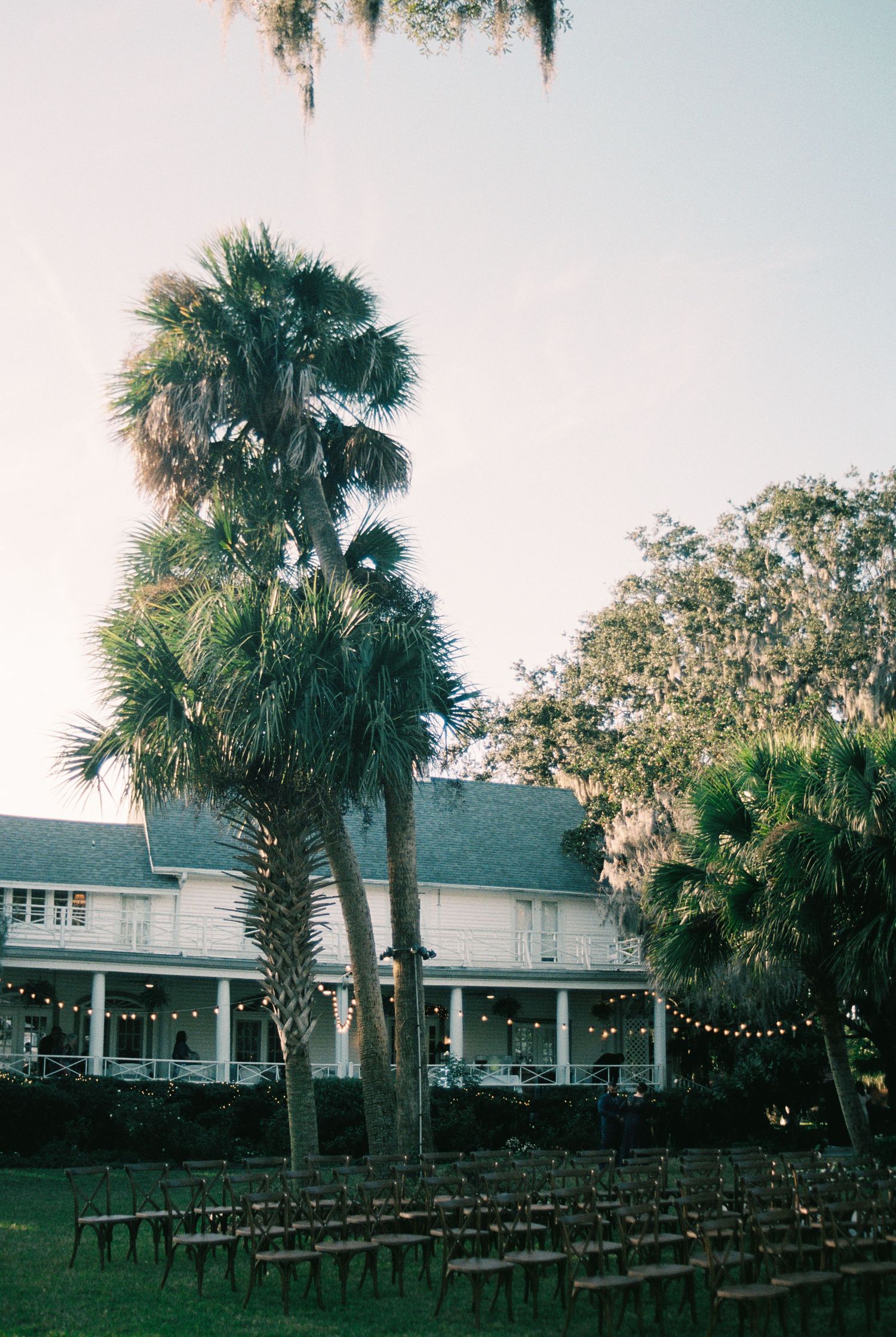 Rows of chairs face a white Southern plantation-style house with palm trees and Spanish moss at sunset.