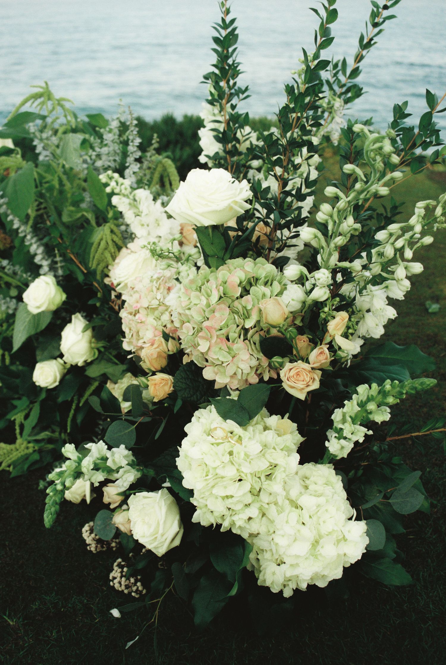 A lush floral arrangement of white hydrangeas and roses with greenery cascading along a waterfront setting.