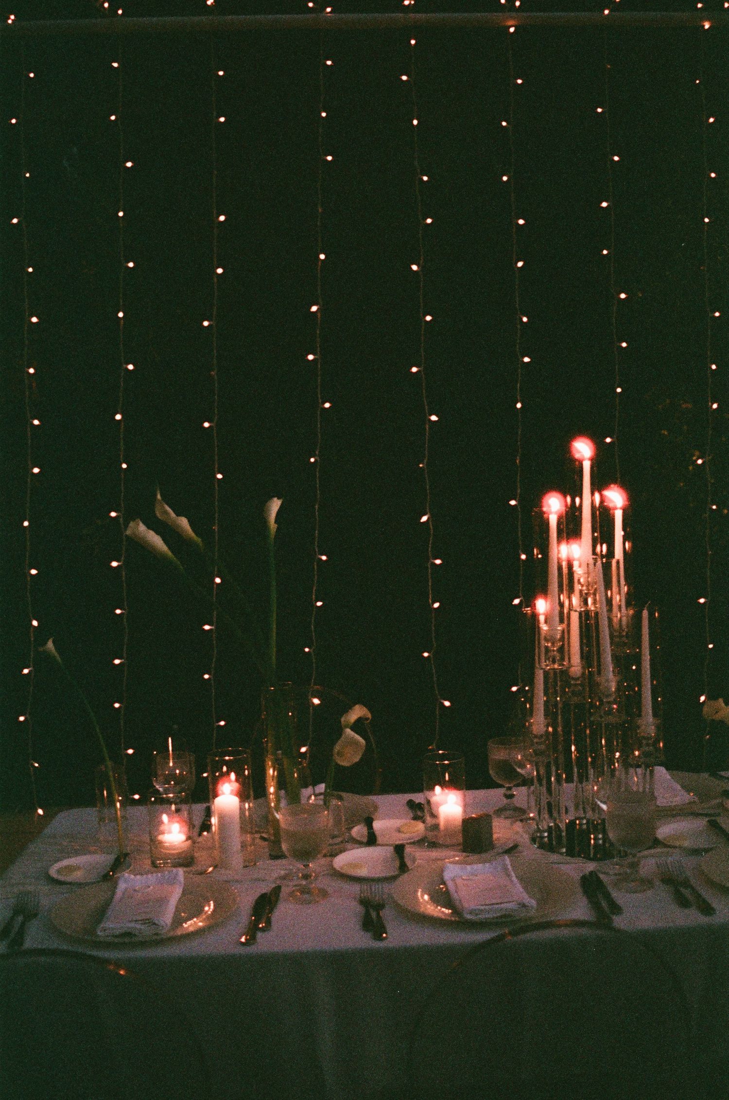 An elegant candlelit table setting with white linens and a backdrop of twinkling string lights creates a romantic ambiance.