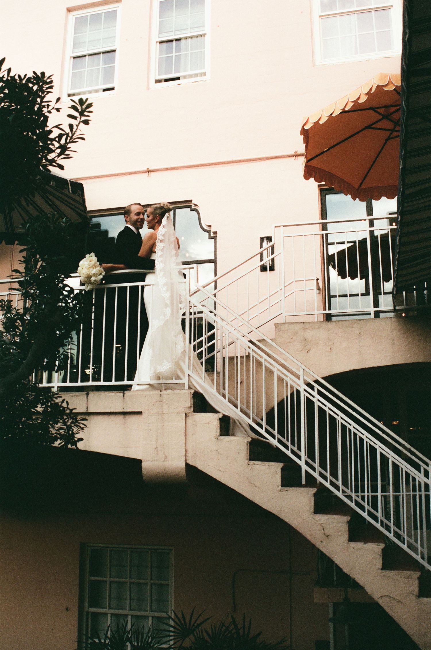 A white wedding dress flows down an outdoor staircase with a terracotta umbrella and cream-colored building in the background.