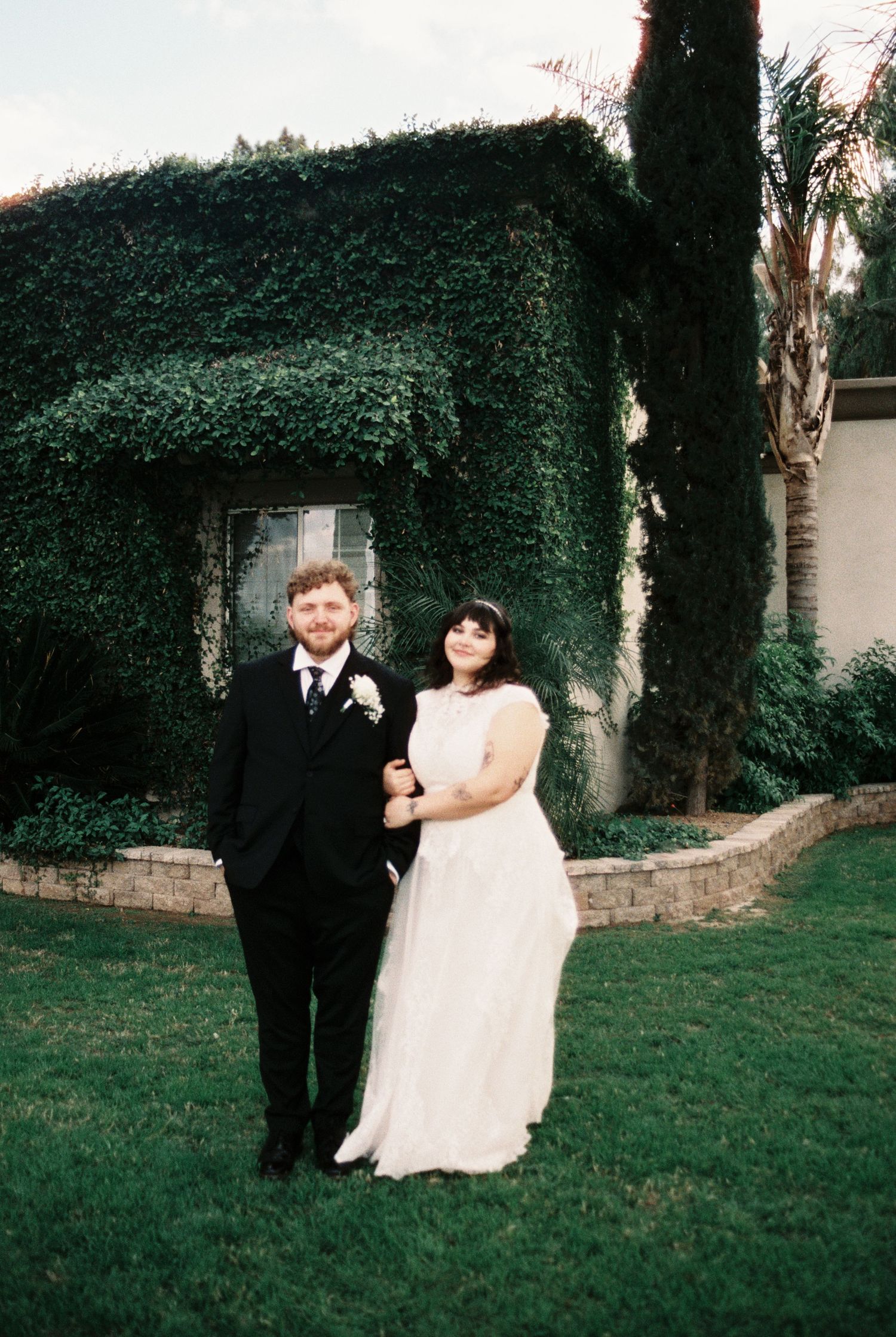 A couple in formal wedding attire poses together in a garden setting with ivy-covered walls during golden hour.