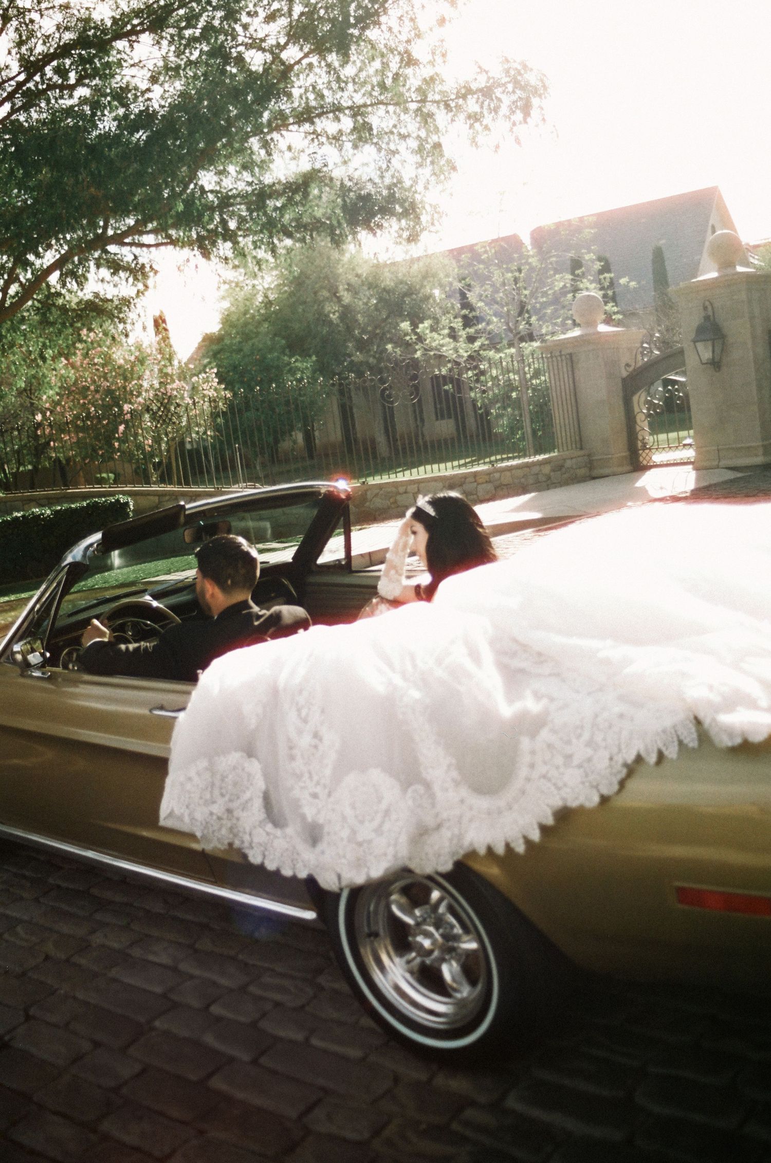 Bride in white lace gown seated in classic gold convertible car with groom, parked outside elegant estate gates.