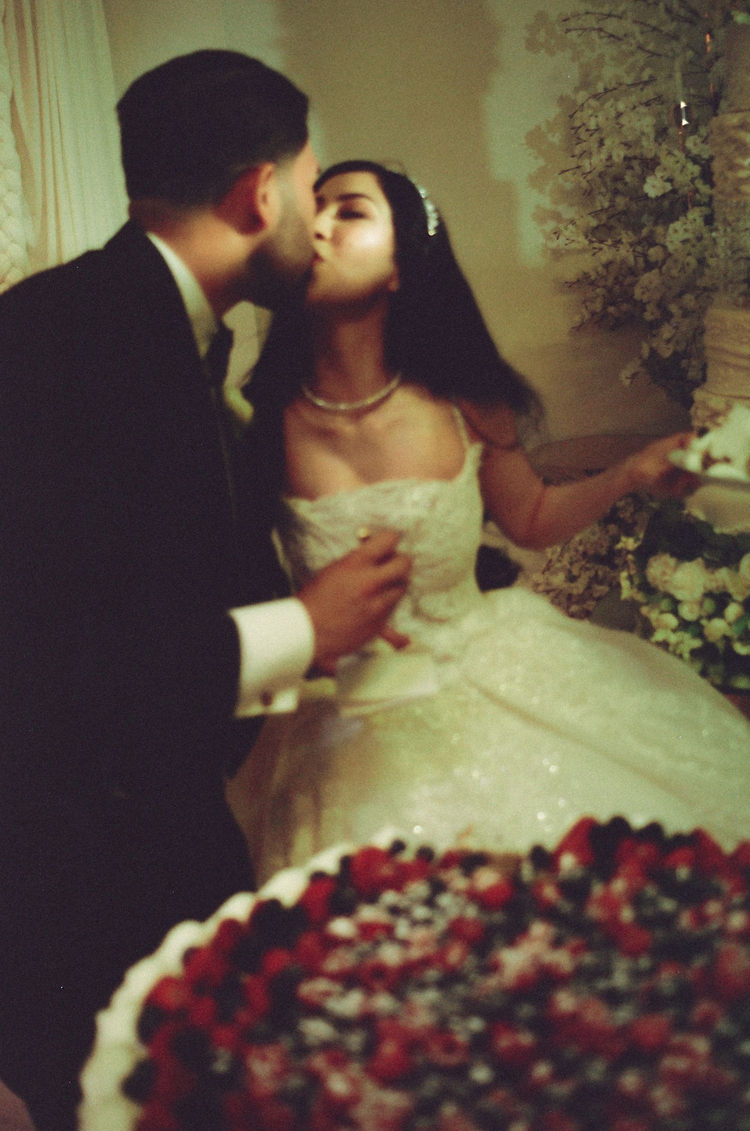 Bride and groom sharing a kiss at their wedding reception with a berry-topped cake in the foreground.