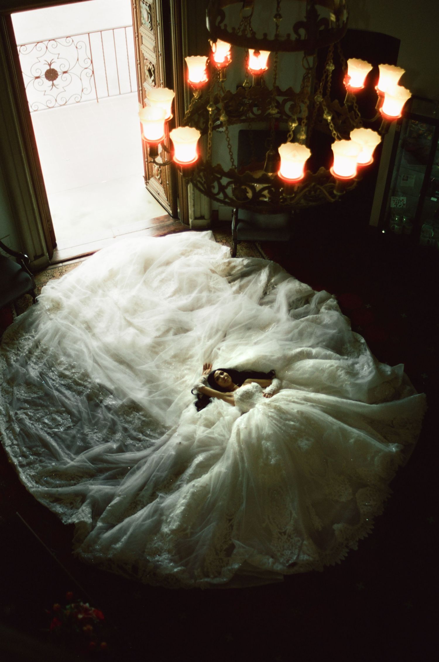 Bride in voluminous white wedding gown lying on floor beneath ornate chandelier with glowing candles in dimly lit room.