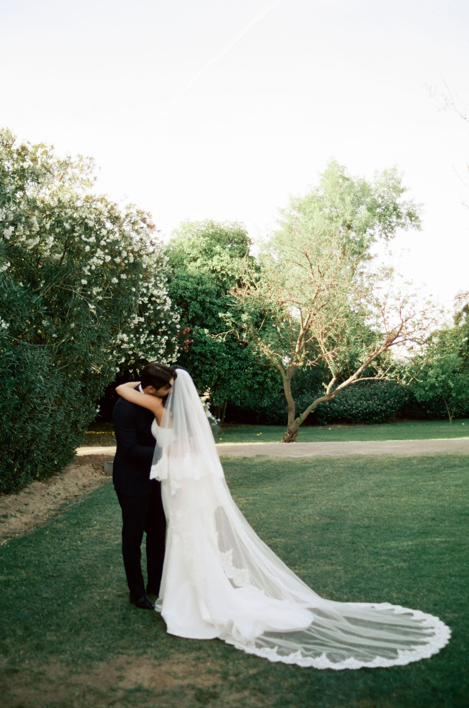 Bride and groom share a romantic kiss on lush green lawn, with bride's long cathedral veil trailing elegantly behind her.