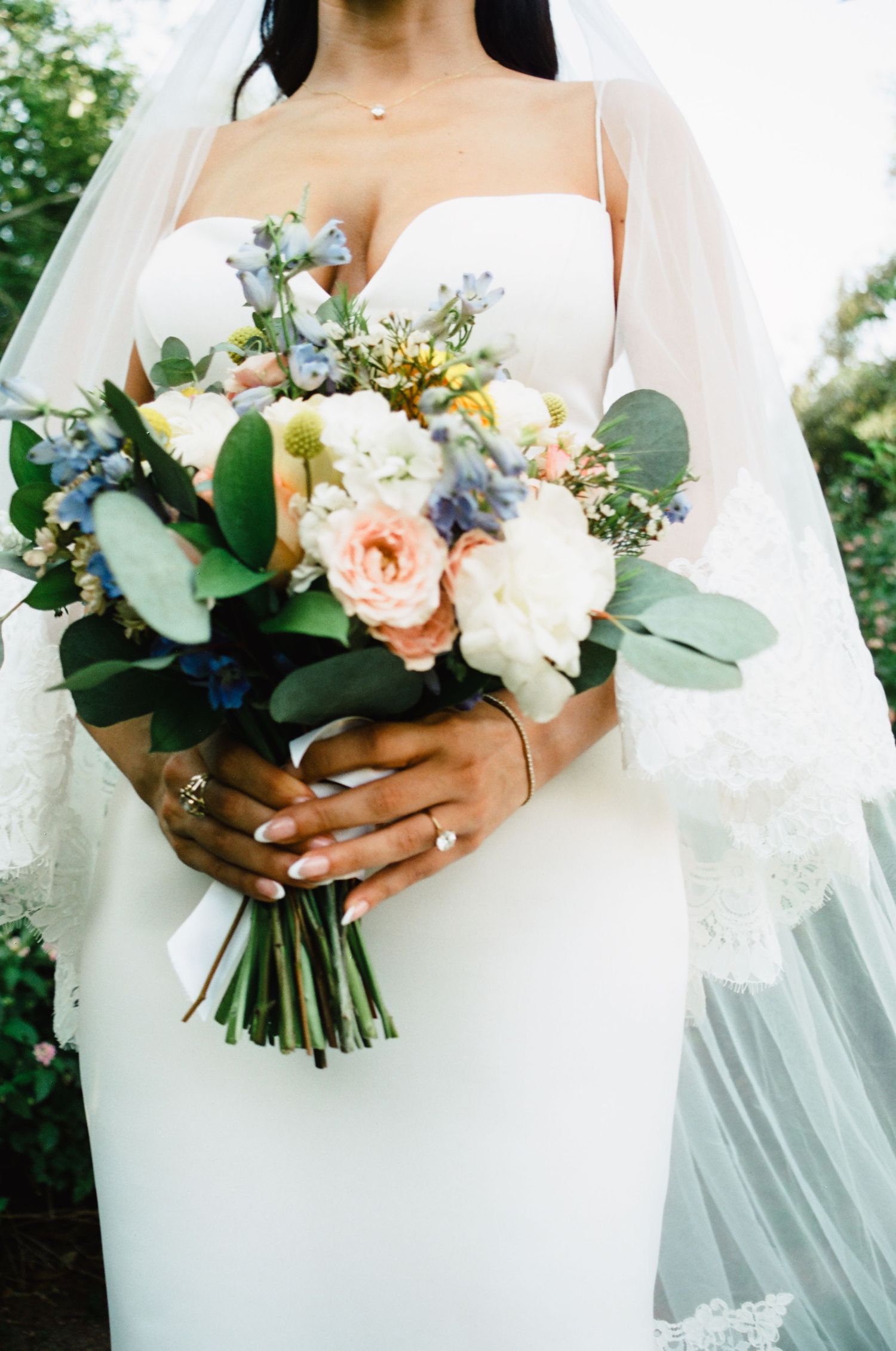 Bride in white gown holding a romantic bouquet of peach roses, white flowers, blue blooms, and eucalyptus with a diamond ring.