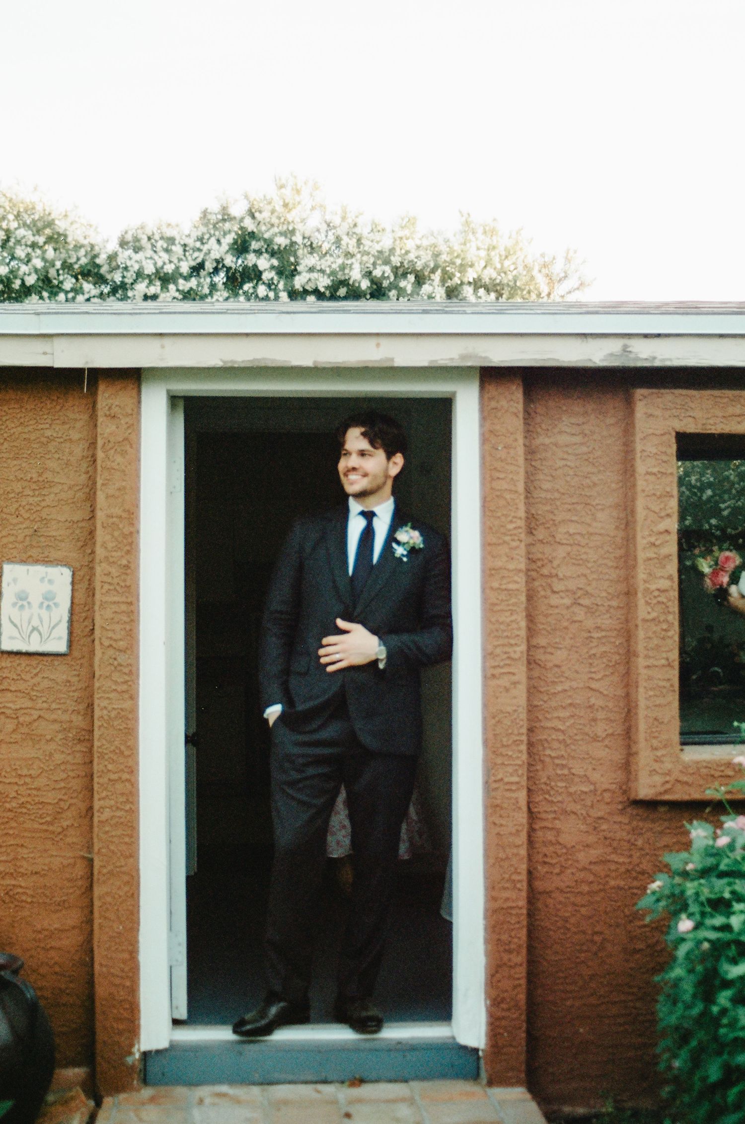 Groom in dark suit with boutonniere standing in doorway smiling on his wedding day.
