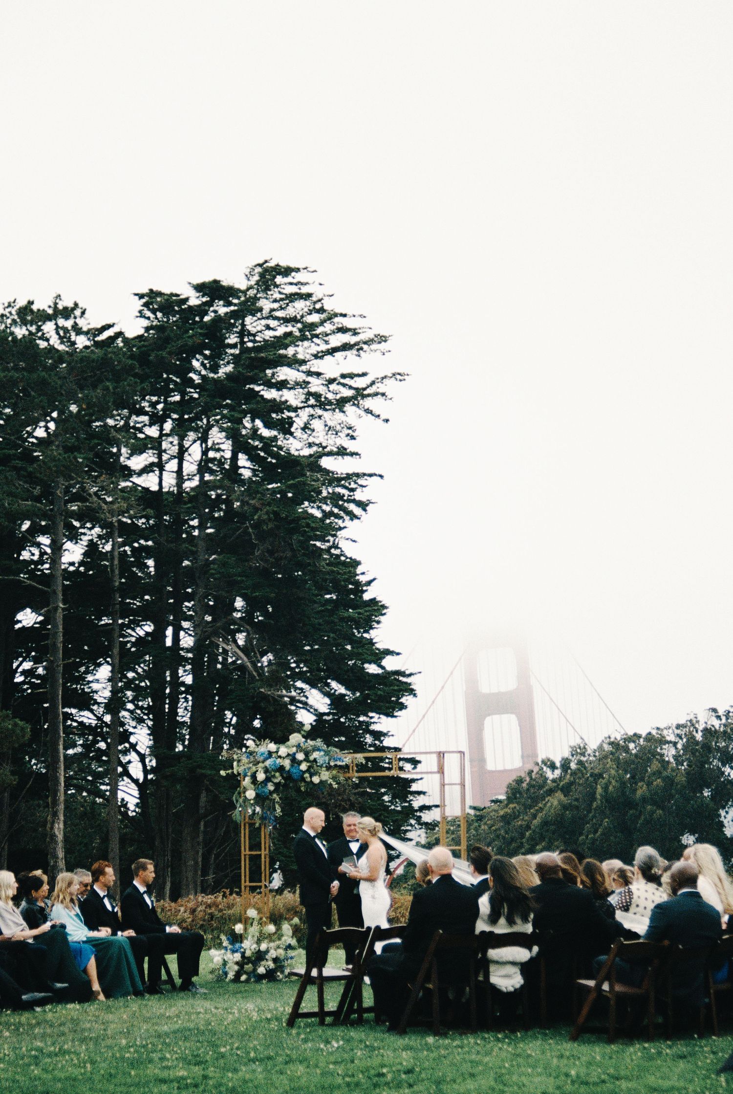 Outdoor wedding ceremony on a lawn with the Golden Gate Bridge visible through fog and tall trees in San Francisco.