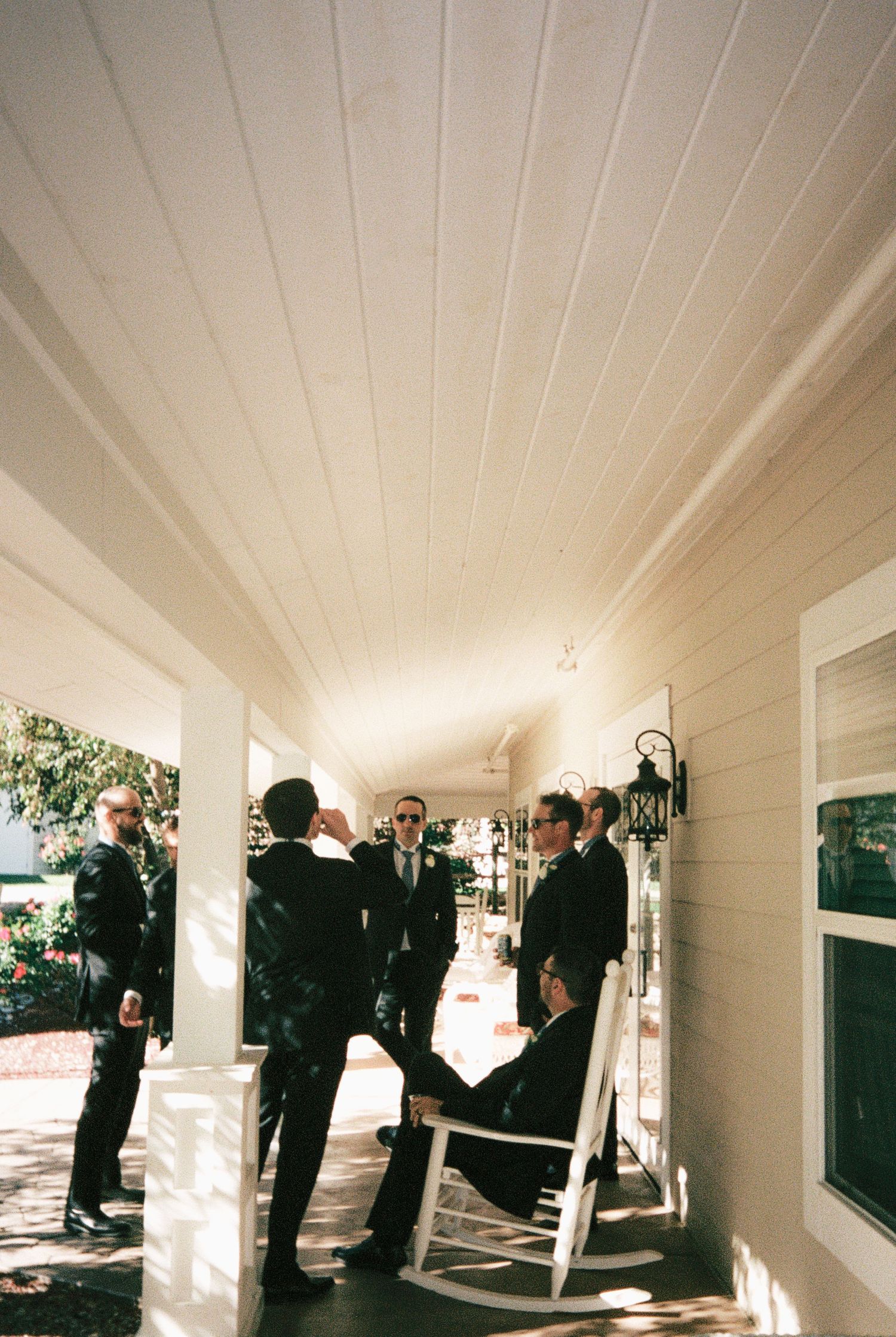 Groomsmen in dark suits gather on a sunlit white porch with rocking chairs before a wedding ceremony.