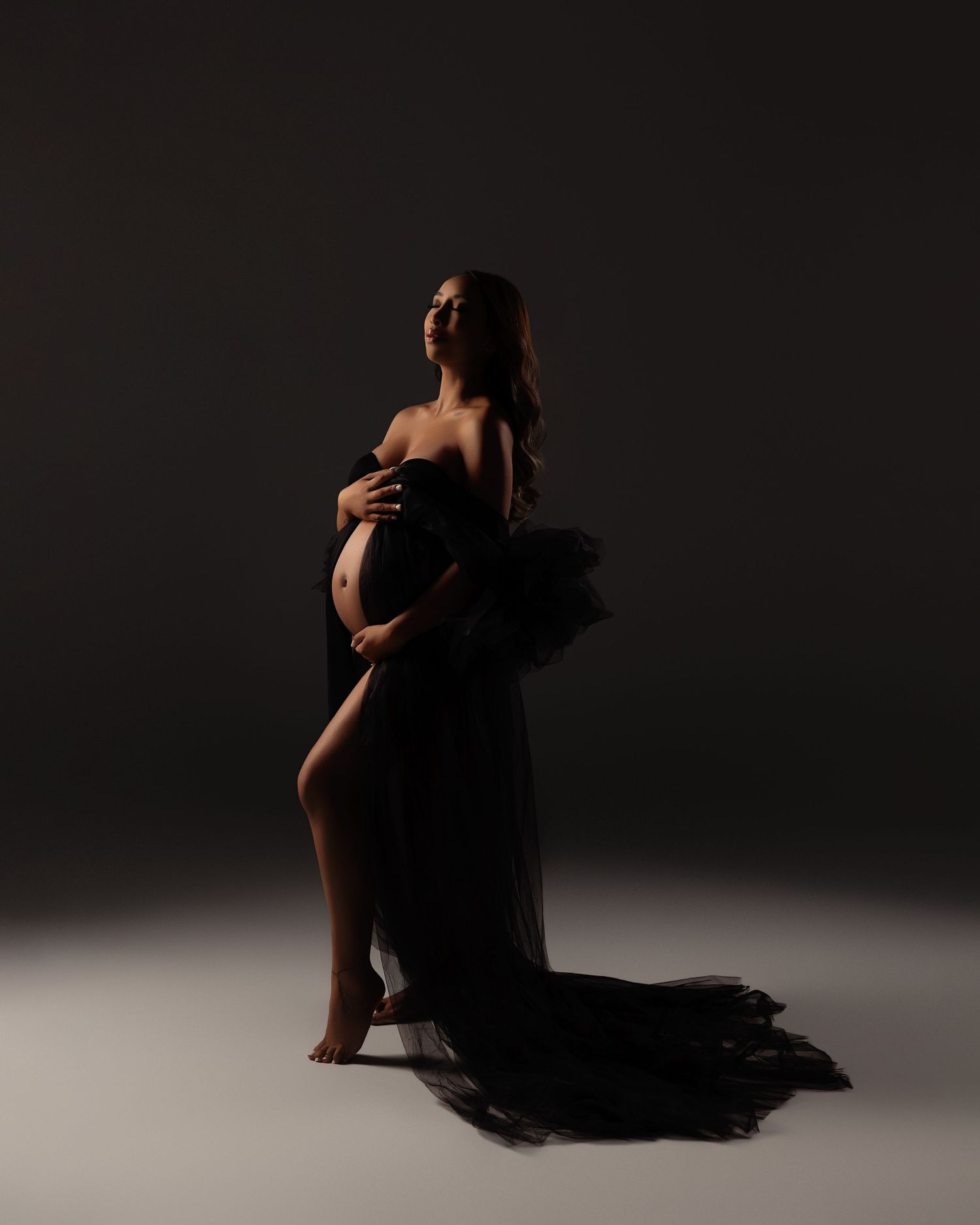 A dramatic silhouette of a pregnant figure draped in flowing black fabric poses against a dark studio backdrop.