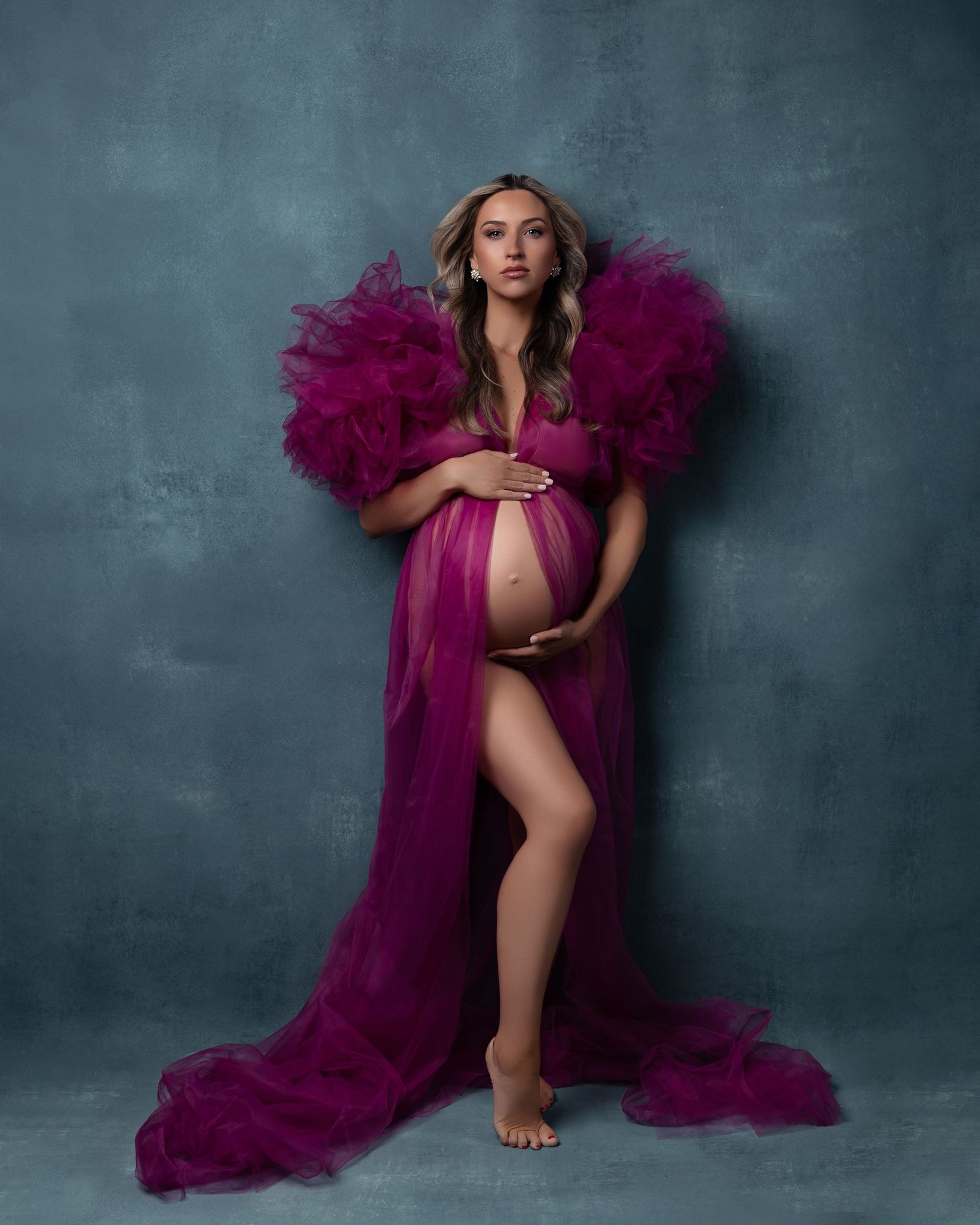 A maternity photo featuring a flowing magenta tulle robe against a moody gray backdrop.