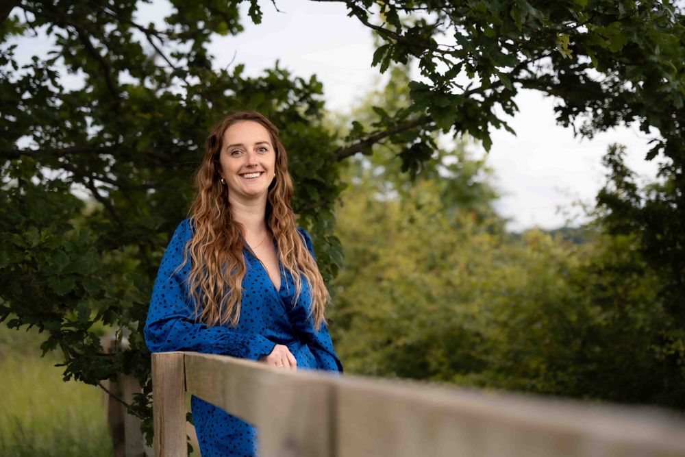 Woman in flowing blue polka dot dress poses by wooden railing in outdoor nature setting. Lincolnshire branding photographer.