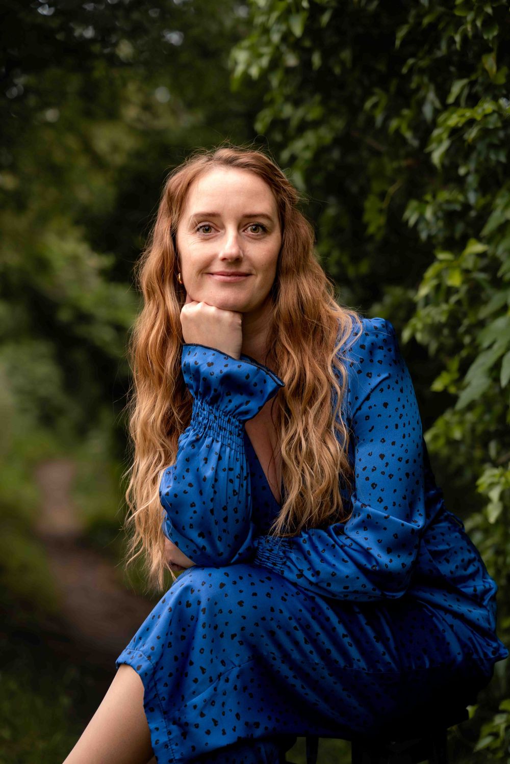 A gentle outdoor portrait showing person in royal blue dress with long wavy hair against green foliage. Lincolnshire branding photographer.
