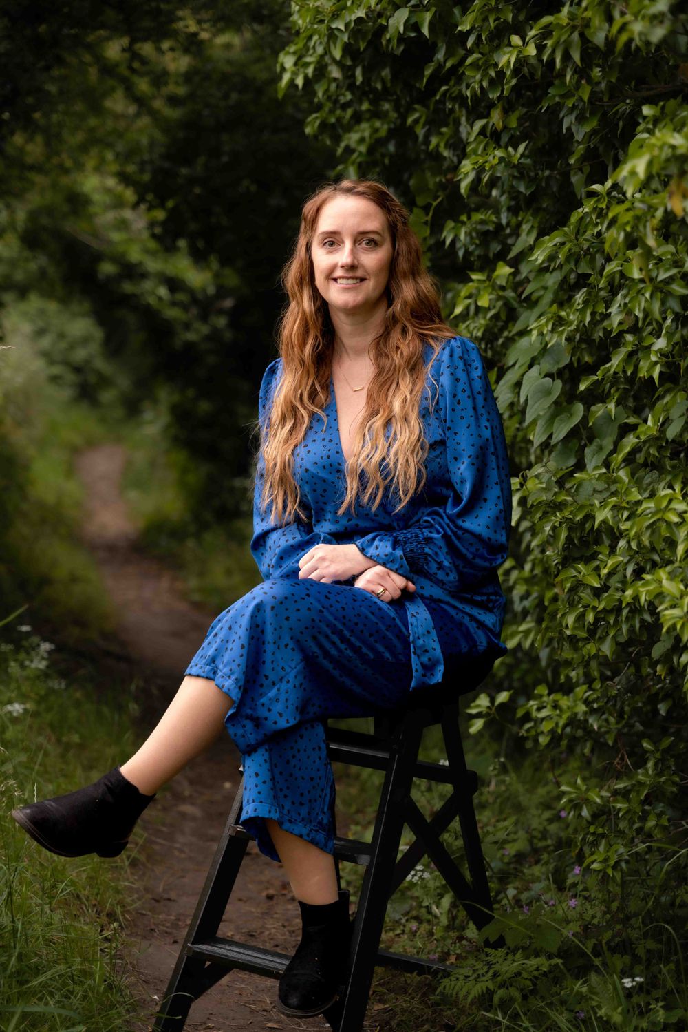 Person wearing blue patterned dress sits on black stool along garden path surrounded by greenery. Lincolnshire branding photographer.