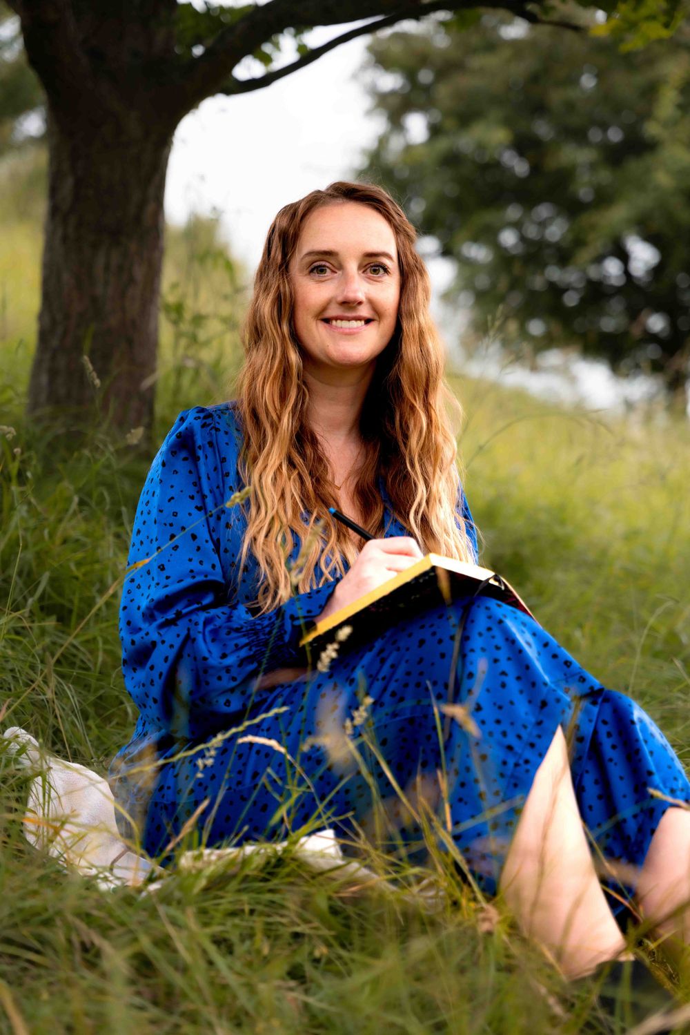 Person in vibrant blue dress sits in tall grass while writing in notebook outdoors. Lincolnshire branding photographer.