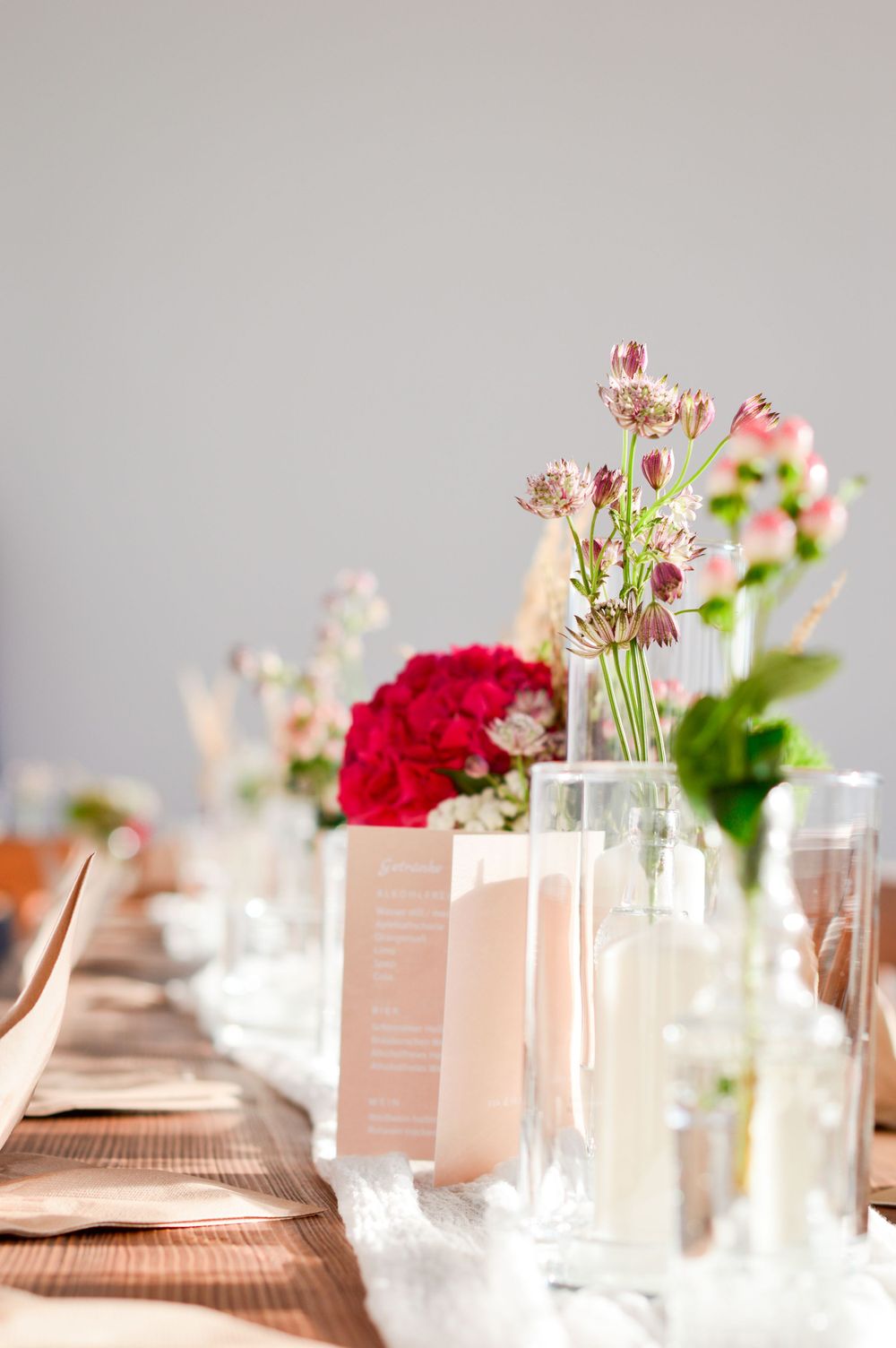 Gedeckter Tisch mit hellbraunen Menükarten und zarten Blumenarrangements in Glasvasen mit roten und rosa Blüten.