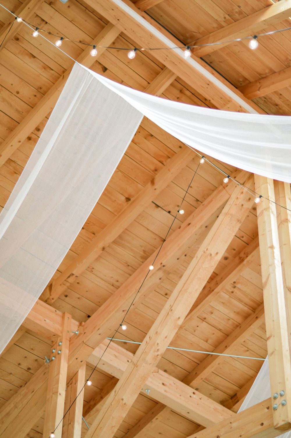 Weißer Stoff, elegant drapiert unter Holzbalken mit Lichterketten an der Decke einer rustikalen Scheune oder eines Veranstaltungsortes.
