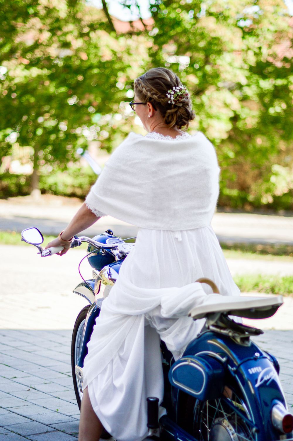 Eine Braut in weißem Hochzeitskleid sitzt an einem sonnigen Tag im Freien auf einem blauen Motorrad.