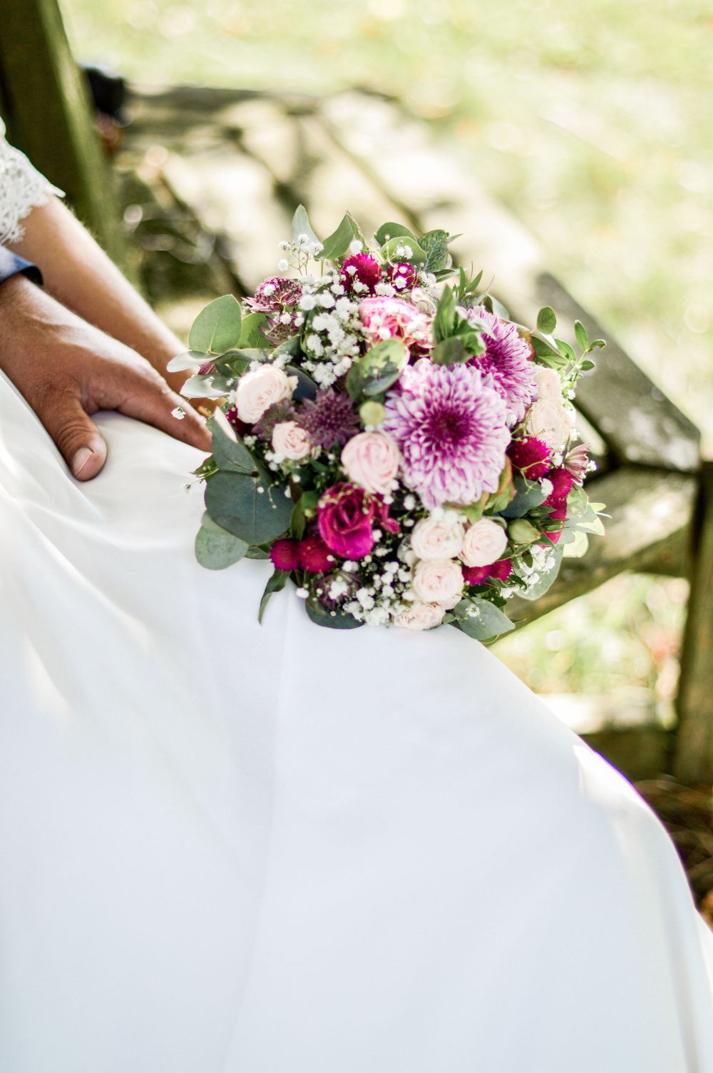 Ein wunderschöner Brautstrauß mit rosa Dahlien, weißen Rosen und grünem Eukalyptus ruht auf einem fließenden weißen Brautkleid.