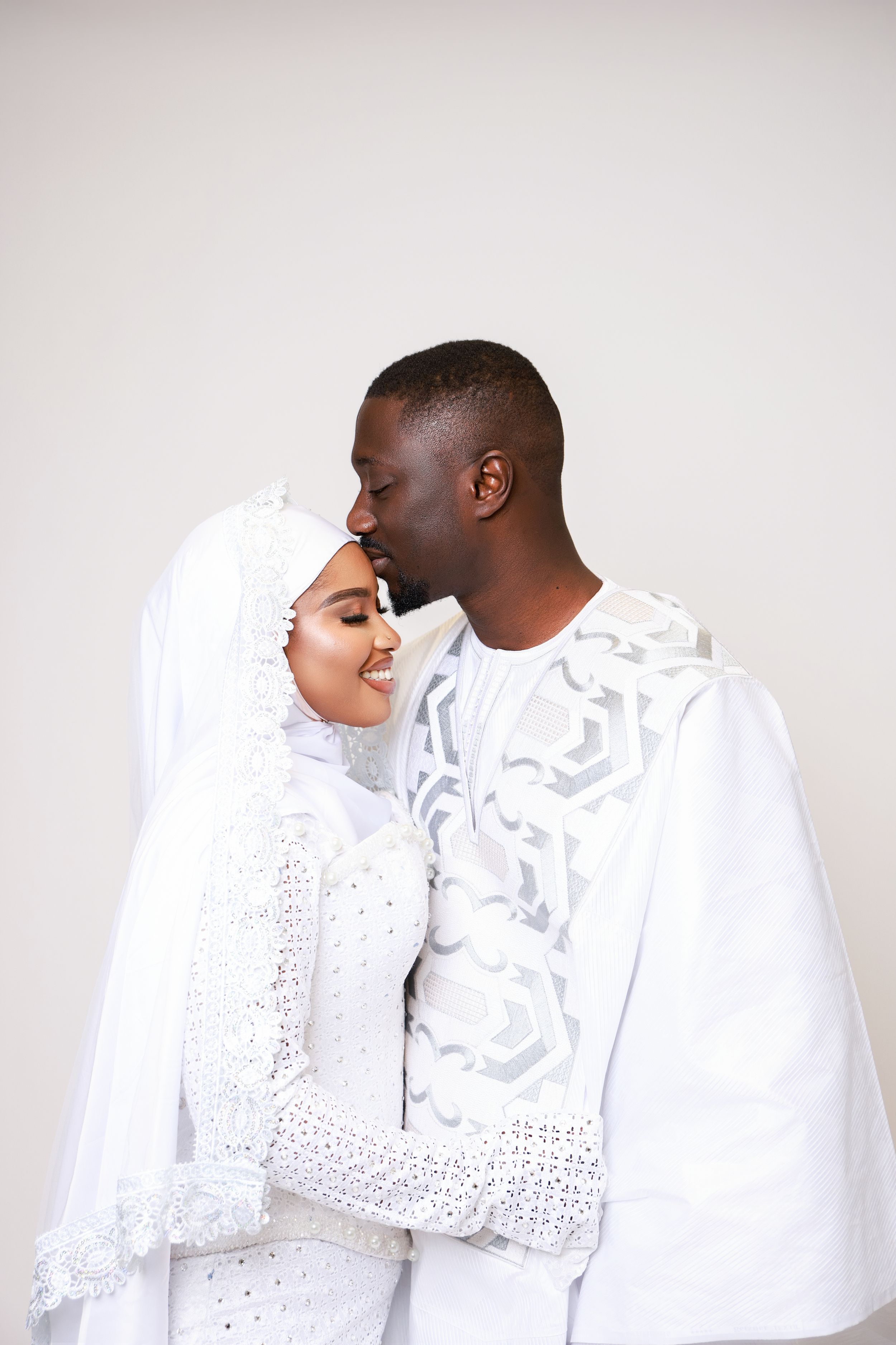 Couple in elegant white outfits share intimate moment against light background.