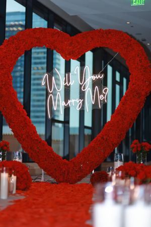 A romantic proposal setup with a large red heart decoration and neon 'Will You Marry Me?' sign illuminated against dark walls.