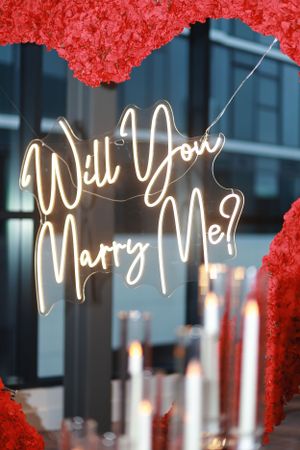 Close-up of glowing white neon text reading 'Will You Marry Me?' against a red heart-shaped backdrop.