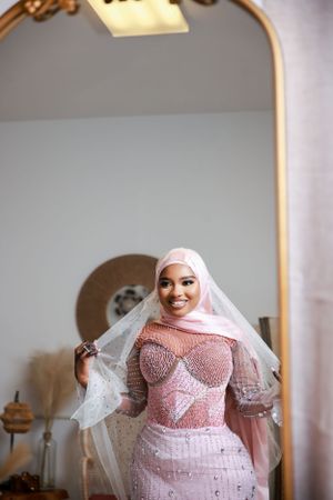 A bride in a pink beaded gown and white veil poses elegantly in front of an ornate gold-framed mirror.