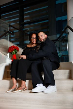 A couple sits together on modern stairs, dressed elegantly with red roses and contrasting formal and casual elements.