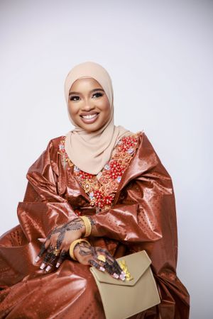 Person wearing traditional copper-colored dress with embroidery and light beige hijab holds envelope while smiling.