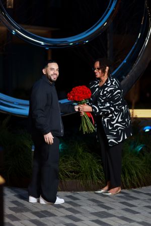 A couple stands beneath a blue circular structure exchanging red roses during a special moment.