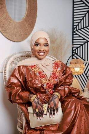 Woman wearing a brown satin dress with red embroidery and a white headscarf poses on a wicker chair.