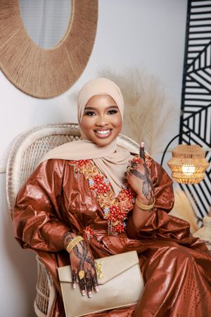 Woman in beige hijab and brown embroidered dress sits in rattan chair with elegant handbag and geometric wall art decor.