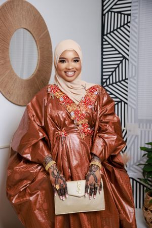 A person in a copper-colored embroidered dress with floral details and a beige hijab poses with a cream handbag.