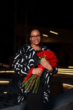 Person in black and white patterned outfit holding vibrant red roses against dark background.