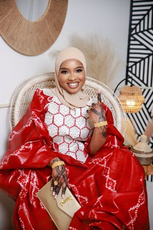 Woman in red and white traditional dress with henna designs seated in wicker chair.