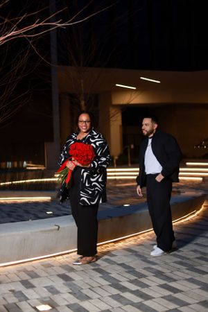 Two people walking in a courtyard, one holding red roses and wearing black and white pattern.