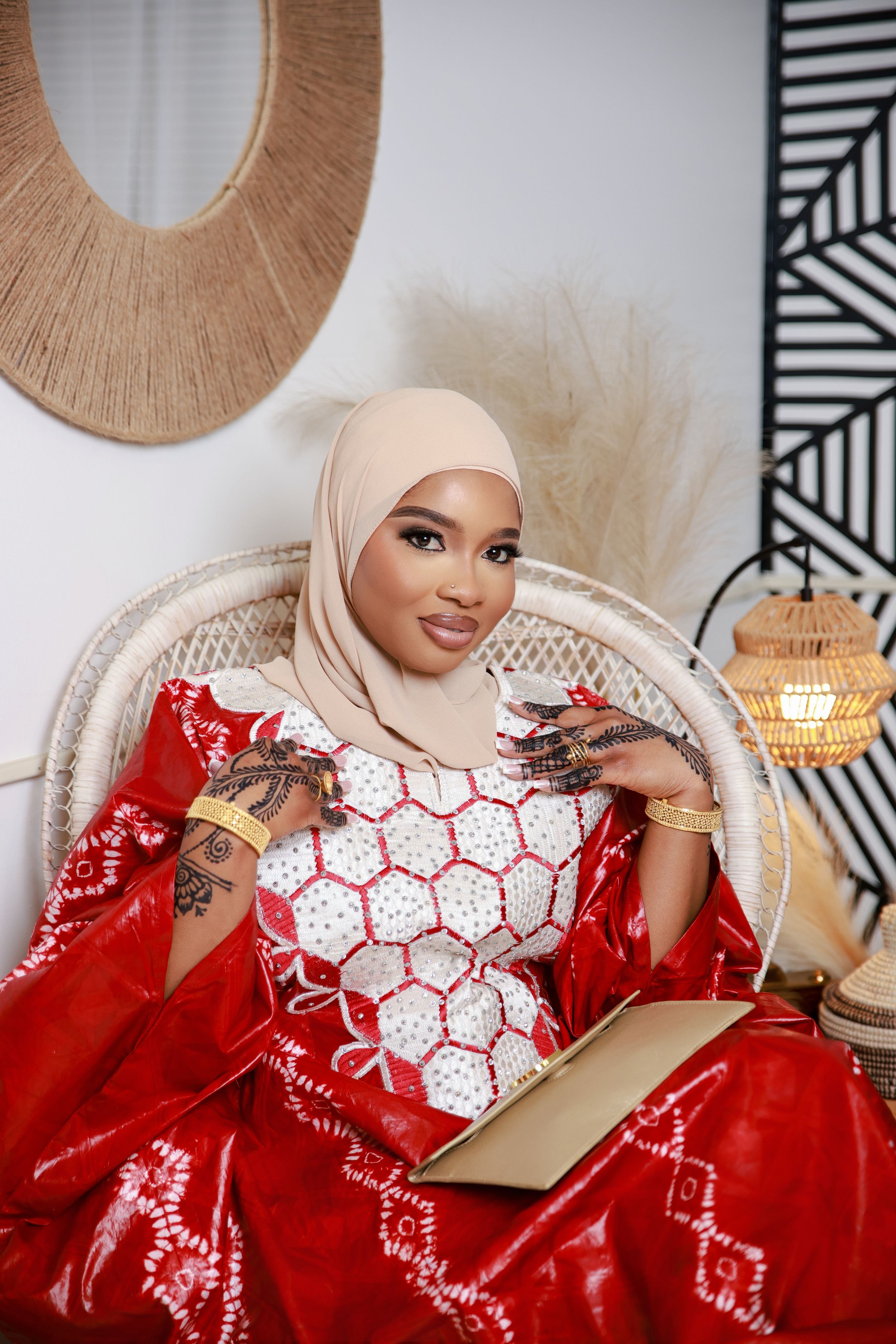 Person in elegant red and white traditional dress with henna designs sits in ornate chair holding book.