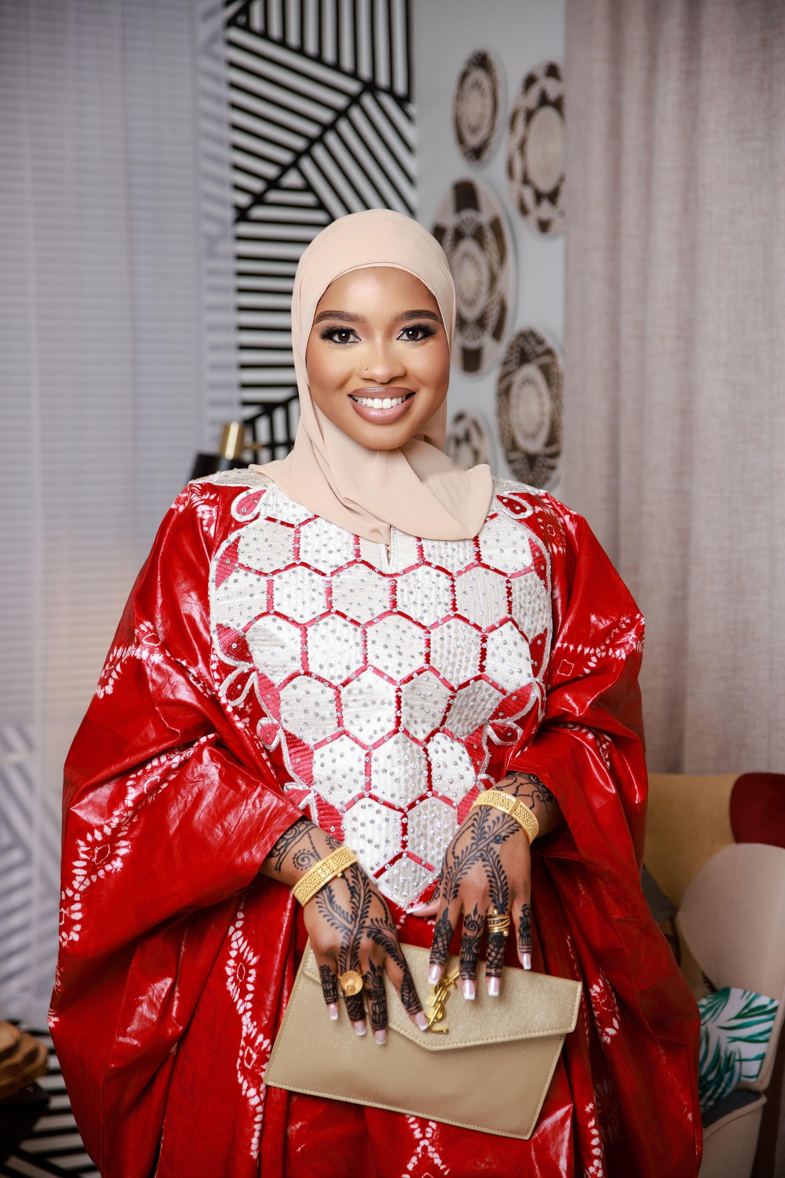 A woman in a vibrant red and white dress with traditional henna designs sits elegantly against a patterned wall.