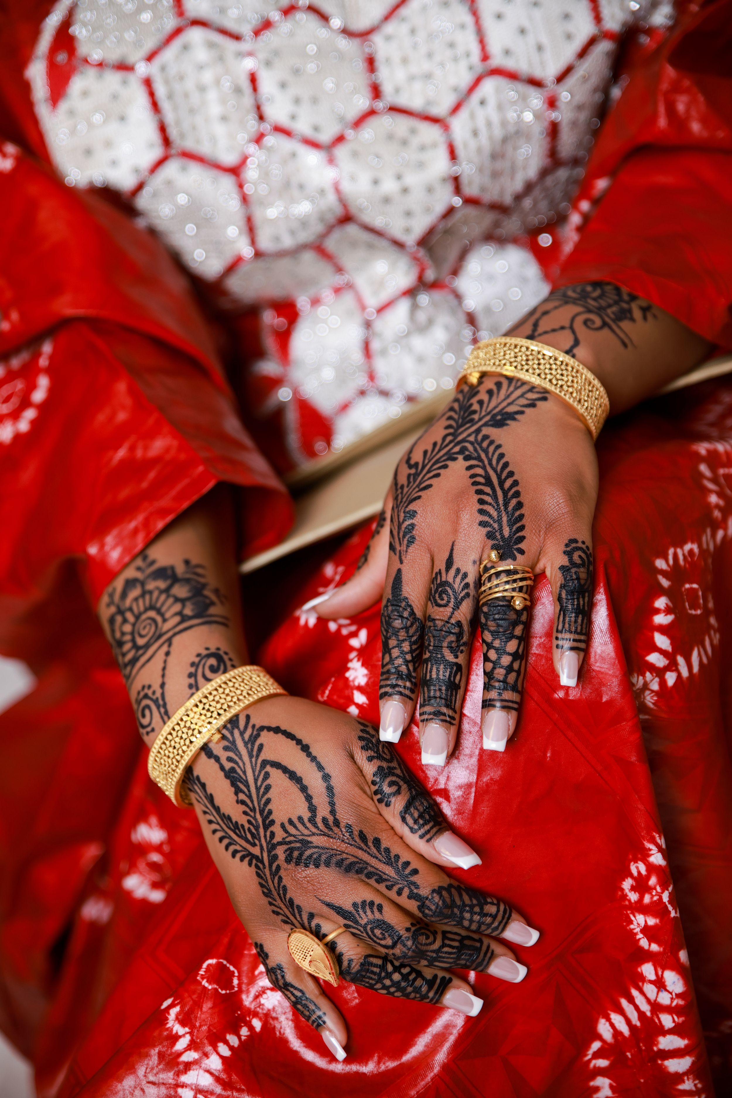 Close-up of hands adorned with intricate henna designs and gold bracelets against a red dress background.