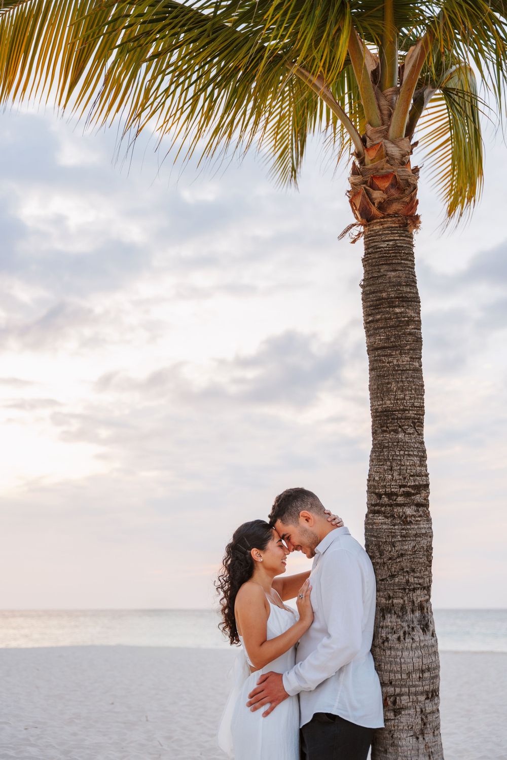 A couple shares an intimate moment under a palm tree on a white sandy beach at sunset.