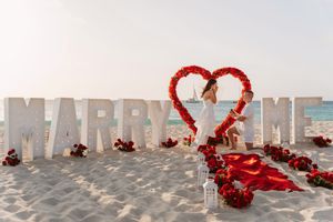 A beach marriage proposal setup with white 'MARRY ME' letters and a heart shape made of red roses in the sand.